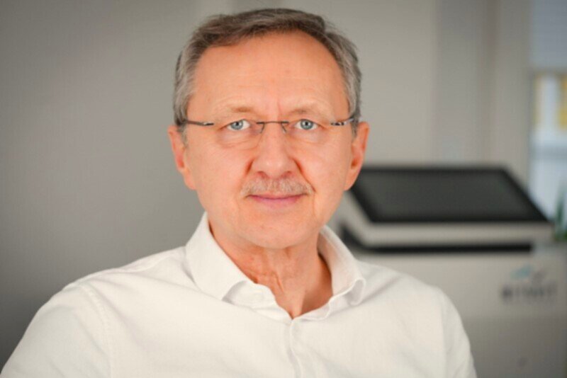 Wolf-Peter Franke, Heilpraktiker, lächelnd in weißem Hemd in seiner Naturheilpraxis, hinter ihm ein IHHT-Gerät.